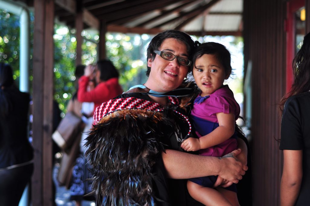 Dayle Takitimu, wearing glasses, stands holding a small child and is wearing ceremonial robes